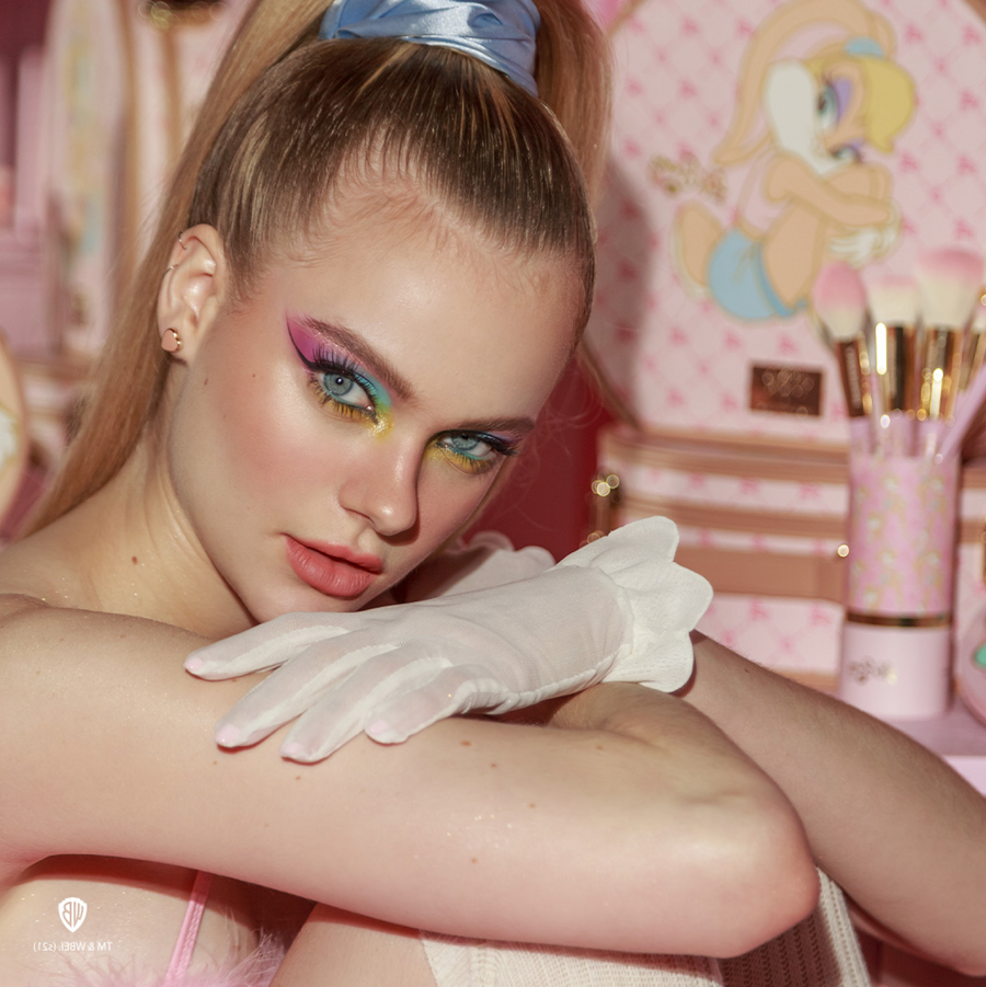 Una persona vestida con suaves colores pastel y guantes blancos está sentada con los brazos cruzados, rodeada de una decoración lúdica y femenina.